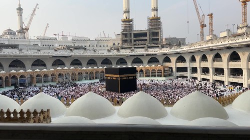A scene of the Grand Mosque in Mecca shows the Kaaba surrounded by crowds of pilgrims and worshippers performing the Tawaf, with the mosque\'s towers and minarets in the background.