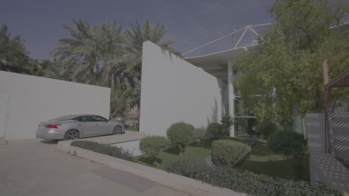 يظهر الفيديو مدخلاً حديثاً لمبنى محاطاً بحديقة خضراء وأشجار نخيل، حيث تبدو العمارة المعاصرة بوضوح مع خطوط بسيطة وهندسة أنيقة، ويظهر موقف للسيارات يحتوي على سيارات مركونة.