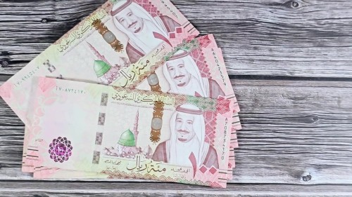 تُظهر الصور مجموعة من الأوراق النقدية السعودية فئة المئة ريال موضوعة بشكل متناسق على سطح خشبي, مع التركيز على التفاصيل الفنية والألوان الموجودة في التصميم.